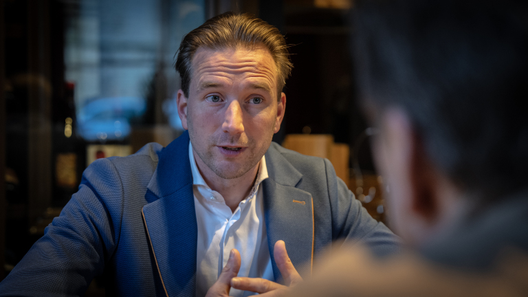 Man in blauw pak in gesprek met ander persoon in een restaurantomgeving.