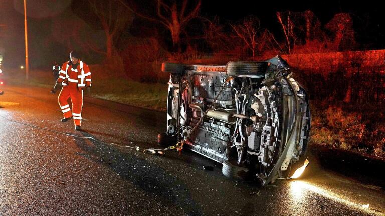 Auto op zijn kant na een ongeval op een donkere weg, met een hulpverlener in reflecterende kleding nabij.