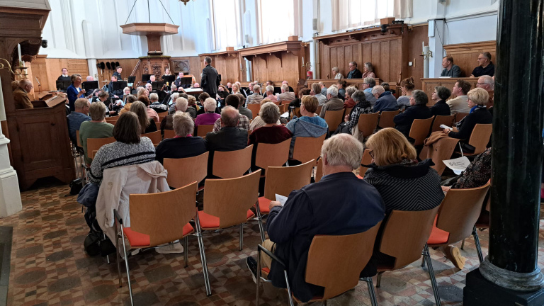 Mensen zitten in een kerk tijdens een bijeenkomst, met een koor en dirigent zichtbaar aan de voorkant.