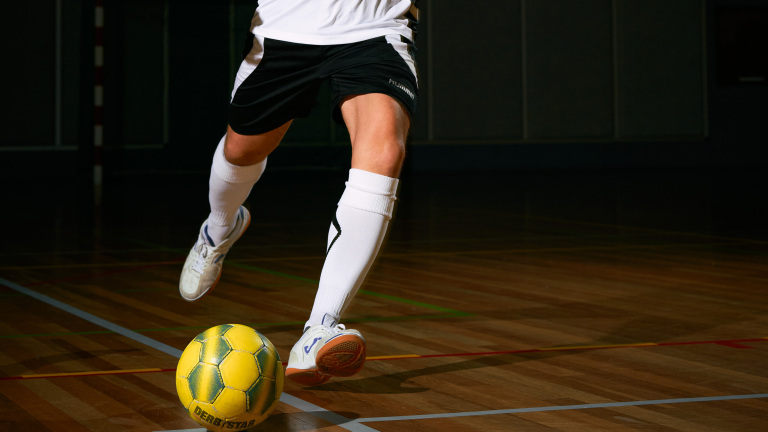 Voetballer dribbelt met een gele bal op een houten zaalvloer.