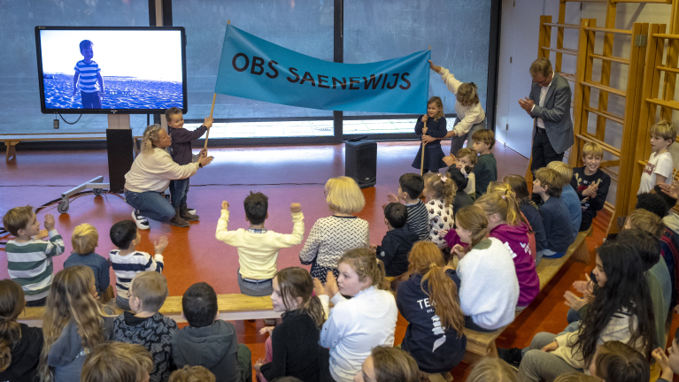 Kinderen in een klaslokaal kijken naar een jongen die een blauw spandoek met de tekst "OBS Saenewijs" vasthoudt.