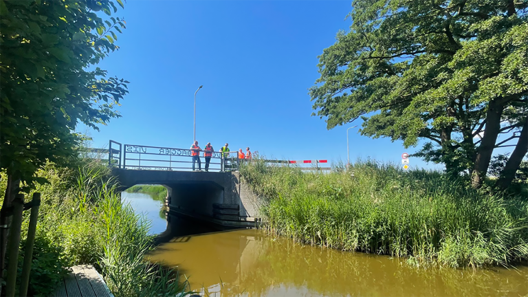 Brug over een kanaal met mensen in felgekleurde werkkleding, omgeven door groen en onder een blauwe hemel.