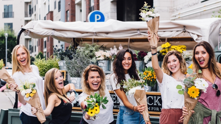 Zes lachende vrouwen met bloemen in hun handen voor een bloemenkraam in een stedelijke omgeving.