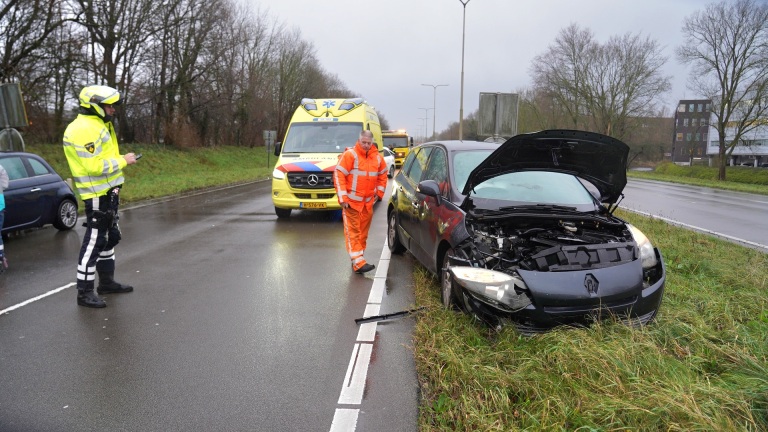 Auto-ongeluk met schade aan een zwarte Renault langs de weg; politieagent en ambulance aanwezig.