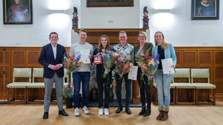 Groep van zes mensen poseert met bloemen en certificaten in een zaal met schilderijen aan de muur.