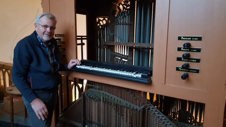 Een man staat naast een ouderwets orgel met zichtbare pijpen en handgrepen voor verschillende registers.