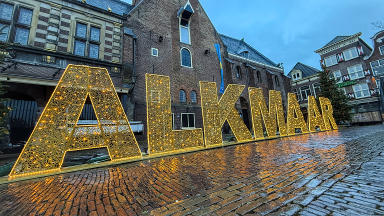 Verlichte gouden letters "ALKMAAR" op een natte klinkerstraat met historische gebouwen op de achtergrond.