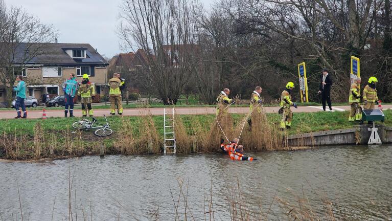 Reddingsteam haalt persoon uit het water, met omstanders en twee fietsen langs de oever.