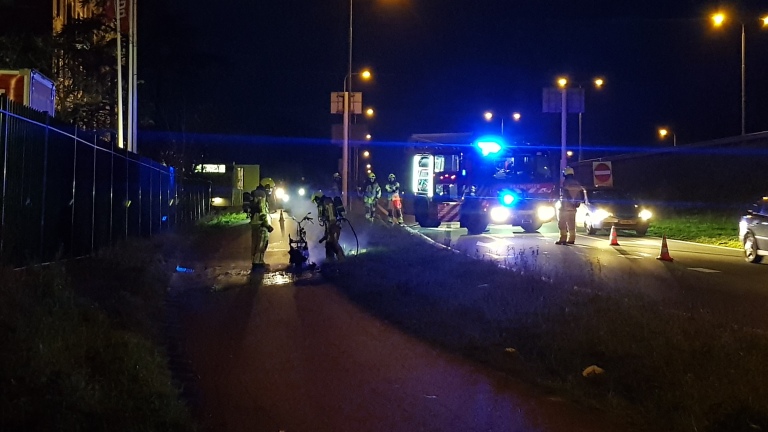 Nachtelijke scène met brandweerlieden die langs de weg een brand blussen, verlicht door blauwe lichten van een brandweerwagen.