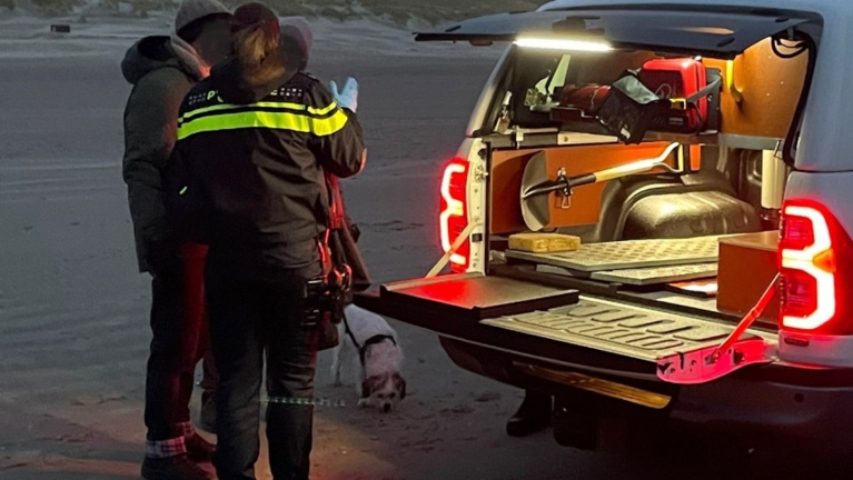 Een politieagent met rug naar de camera in gesprek met twee personen bij een open achterbak van een voertuig op een strand.