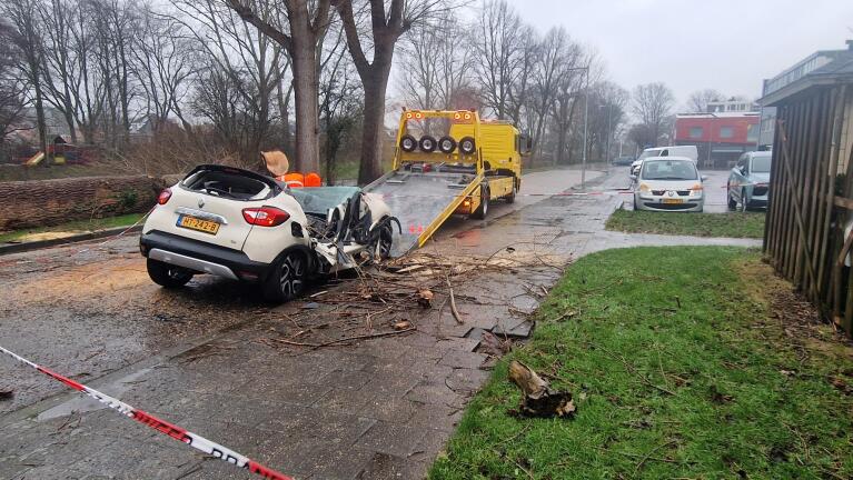 Auto zwaar beschadigd door omgevallen boom, met een sleepwagen die het voertuig oplaadt. Op de achtergrond zijn bouwhekken met rood-wit lint zichtbaar.