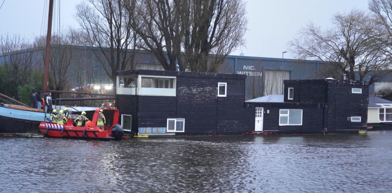 Een woonboot met brandweerboot en brandweerlieden in een kanaal, met een loods op de achtergrond.