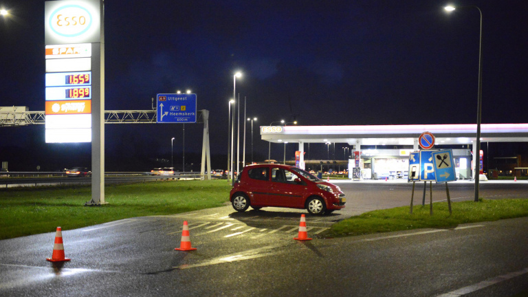 Rode auto geparkeerd bij een Esso tankstation langs een snelweg 's nachts, met verkeersborden en gele verkeerskegels op de voorgrond.