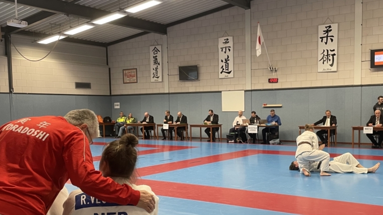 Judo wedstrijd in een dojo met toeschouwers op een bank, een coach in rood en judokas in wit op de mat.