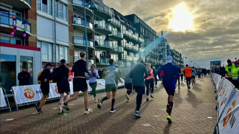 Hardlopers op een promenade tijdens een marathon, met omstanders langs de kant en een bewolkte lucht met zon op de achtergrond.