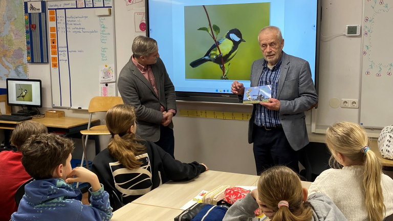 Twee mannen geven een presentatie in een klaslokaal, omringd door leerlingen die aandachtig luisteren. Op het scherm staat een afbeelding van een vogel.