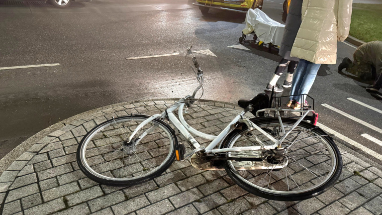 Een omgevallen witte fiets op een trottoir bij een weg met enkele mensen en een ambulance op de achtergrond.