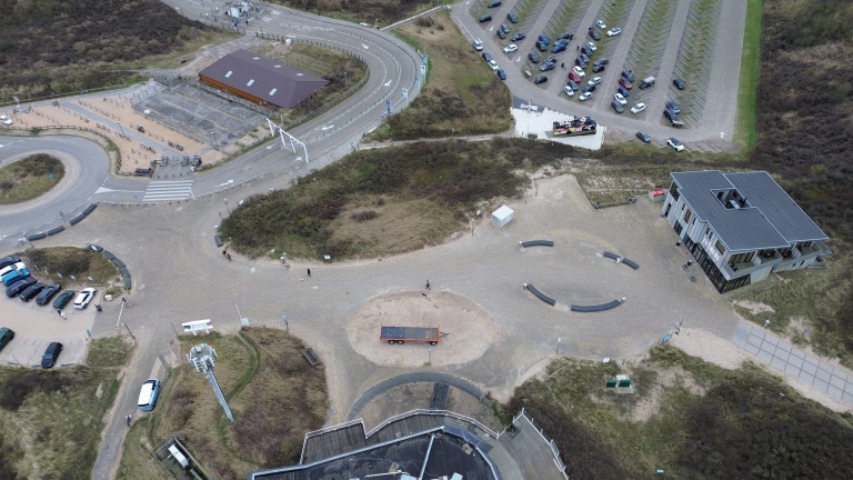 Luchtfoto van een parkeerterrein en gebouwen omgeven door duinen en wegen.