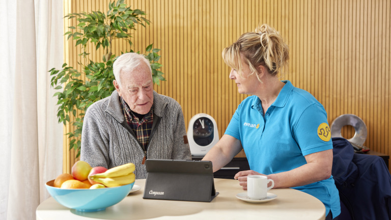 Oudere man en zorgverlener in gesprek aan tafel, met een tablet voor hen en een schaal met fruit.