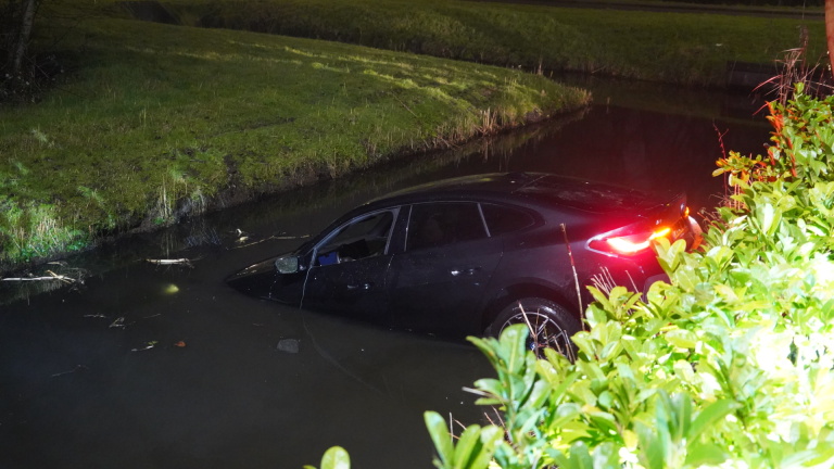 Auto deels ondergedompeld in een sloot, met rode achterlichten aan en omringd door gras en struiken.