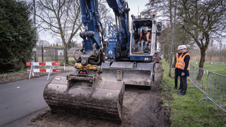 Graafmachine aan het werk langs een weg met twee mensen in bouwhelmen en veiligheidsvesten die toekijken.