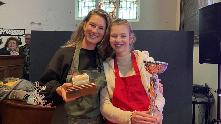 Twee vrouwen poseren in een kerk met trofeeën en schorten van "Heel Schermerhorn Bakt".