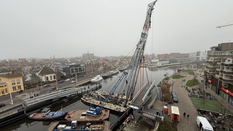 Een hijskraan tilt een brugdeel over een kanaal in een stedelijk gebied met huizen en boten op de achtergrond onder een mistige hemel.