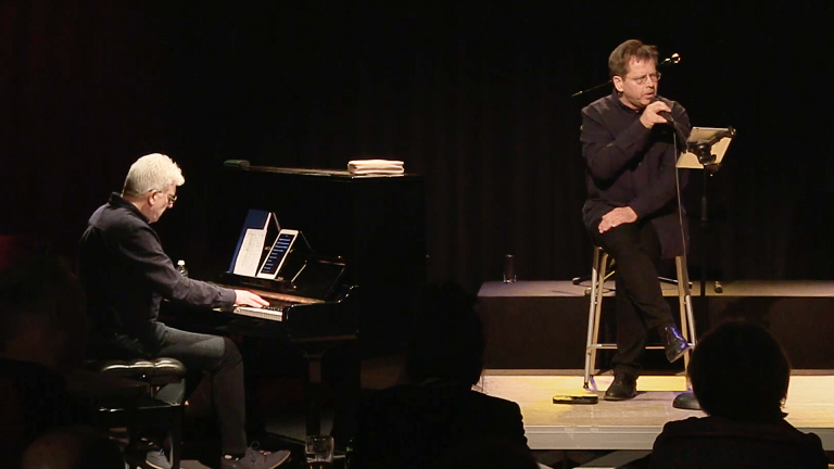 Twee mannen op een podium; de ene speelt piano, de andere zingt in een microfoon.