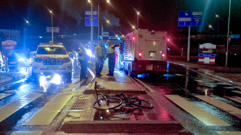 Een verkeersongeval 's nachts met een beschadigde fiets op een zebrapad, omringd door politievoertuigen en verkeersmedewerkers op een natte weg.