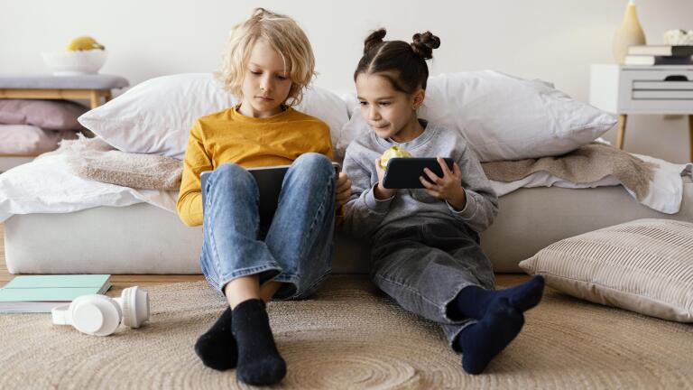 Twee kinderen zitten op de vloer met een tablet en smartphone, in een gezellige kamer met kussens en boeken.