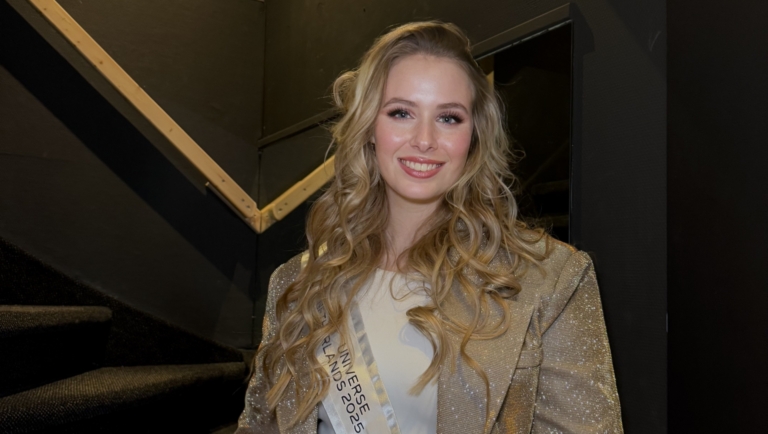 Een vrouw met lang, blond haar draagt een gouden glinsterende outfit en een sjerp met de tekst "Finalist Noord-Holland Miss Universe Netherlands 2025" bij een trap.