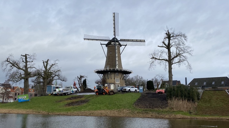 Windmolen naast water met geparkeerde voertuigen op de voorgrond en bewolkte lucht.