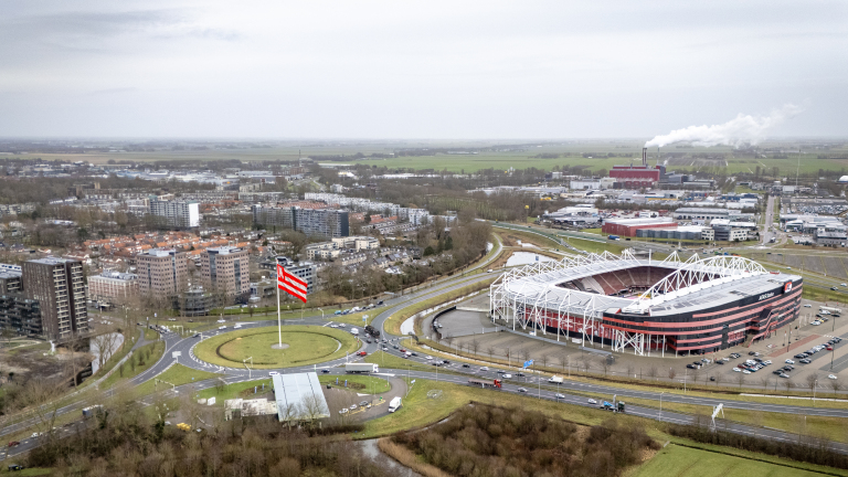 Luchtfoto van een stadion met een groot rood-wit gestreepte vlag op een rotonde, omringd door wegen, gebouwen en een weids landschap.