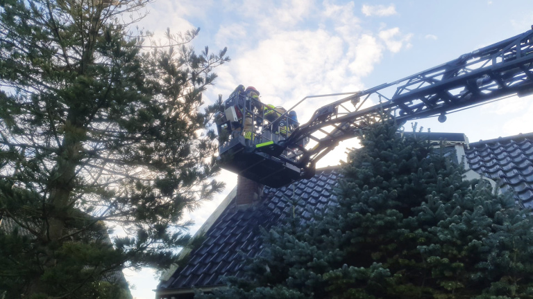 Brandweerlieden op een hoogwerker bij een huisdak omringd door bomen.