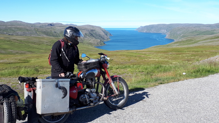 Persoon op een motorfiets met zijspan, met een helm op, staande op een weg, met uitzicht op een groene heuvelachtige kust en blauwe zee.