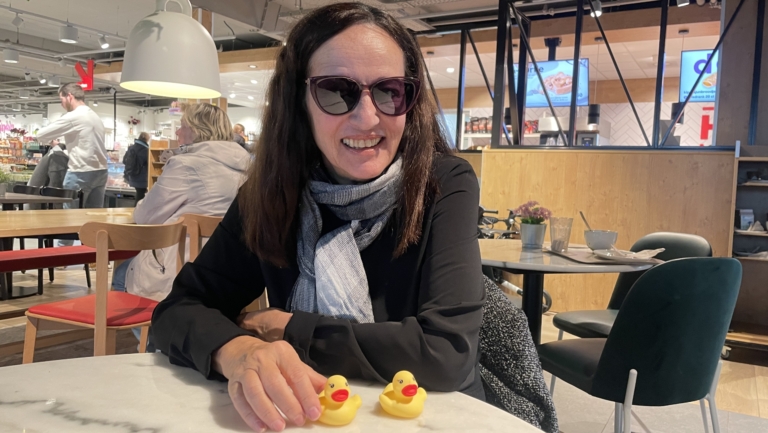 Een vrouw met zonnebril zit in een café en houdt twee gele badeendjes vast op een marmeren tafel.