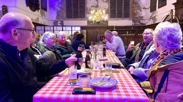 Mensen zitten aan een lange tafel met rood-wit geblokt tafelkleed in een oude kerkzaal, gezellig pratend en drinkend.