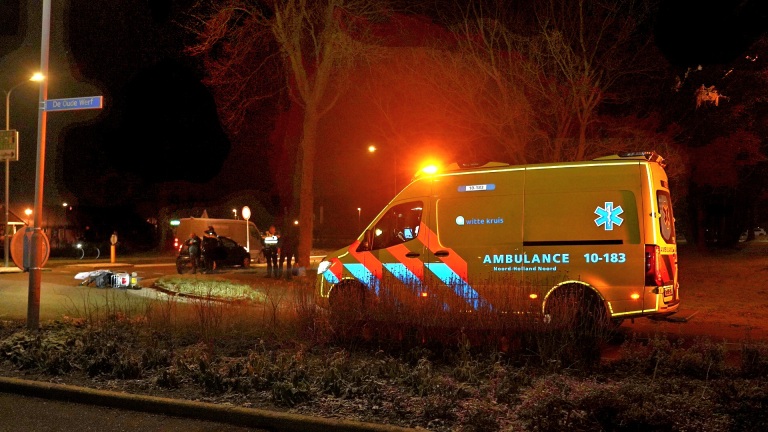 Een ambulance staat geparkeerd langs een donkere straat met zwaailichten aan; twee mensen staan naast een auto op de achtergrond.