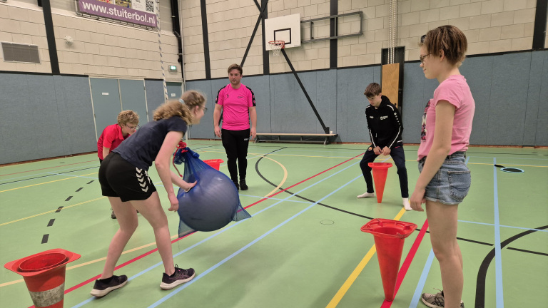 Een groep mensen in een sportzaal bezig met een spel waarbij een grote bal en kegels centraal staan.