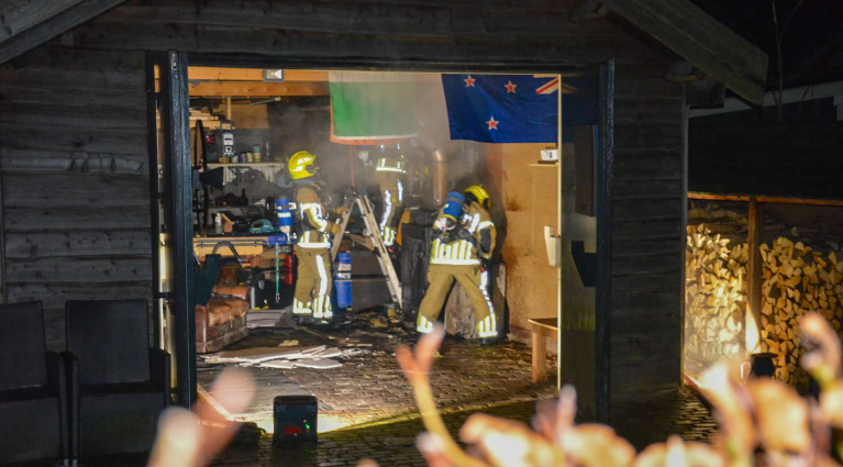 Brandweerlieden blussen een brand in een schuur met rookontwikkeling en vlaggen aan de muur.