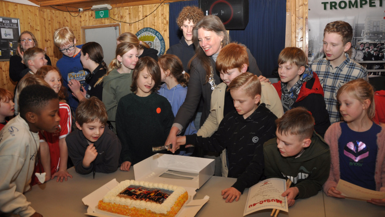 Groep kinderen en een volwassene staan rondom een taart. De volwassene helpt een kind de taart te snijden.