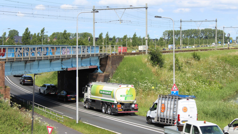 Een vrachtwagen rijdt onder een spoorbrug met graffiti door, omgeven door groene natuur en strakblauwe lucht.