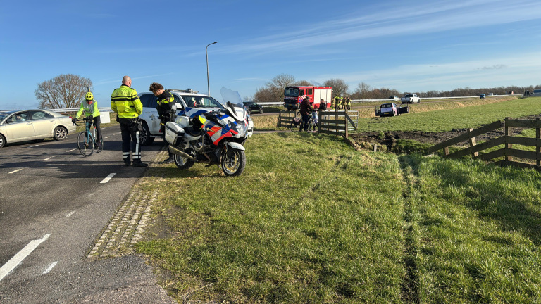 Politie en hulpdiensten op de weg, met een politieagent op een motorfiets en een ander bij een politieauto naast een fietser. Brandweer en een auto in een weiland op de achtergrond.