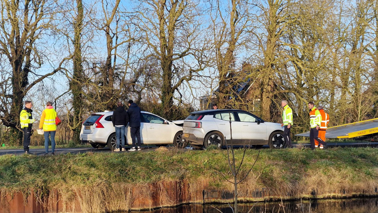 Twee auto's betrokken bij een aanrijding langs een weg met kale bomen; mensen in gele hesjes staan erbij.