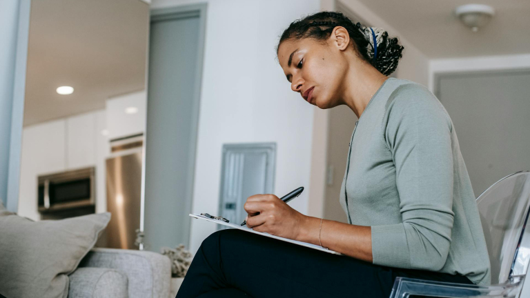 Vrouw zittend en schrijvend op een klembord in een moderne woonkamer.