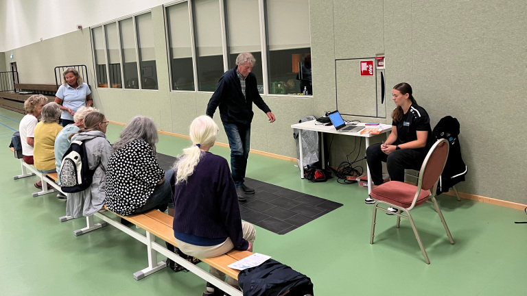 Groep mensen op een bank in een sporthal kijkt naar een persoon die een balansapparaat gebruikt, onder begeleiding van een coach.