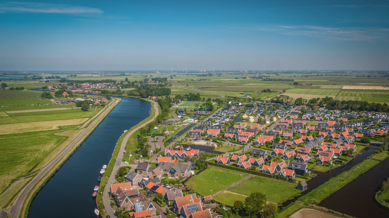 Luchtfoto van een typisch Nederlands landschap met een kanaal, groene velden, en een woonwijk met rode daken.