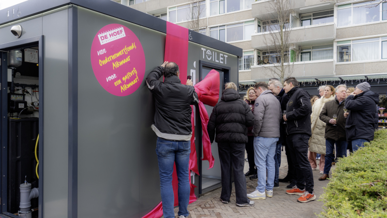 Grijze toiletunit met roze strik wordt onthuld door groep mensen, met geparkeerde flatgebouwen op de achtergrond.