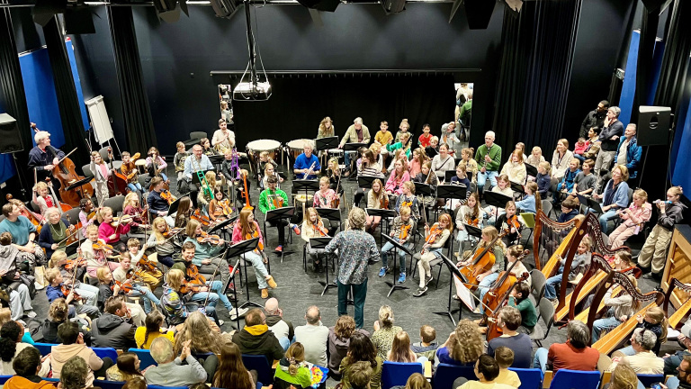 Orkest van kinderen en volwassenen in een zaal met toeschouwers, verschillende instrumenten zoals violen, trombones en harpen zichtbaar.