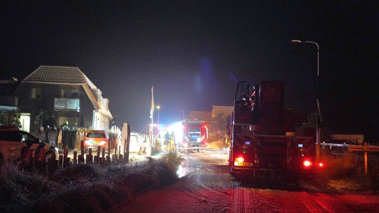 Brandweerwagens en politievoertuigen bij een huis in de nacht, met mensen en felle verlichting.